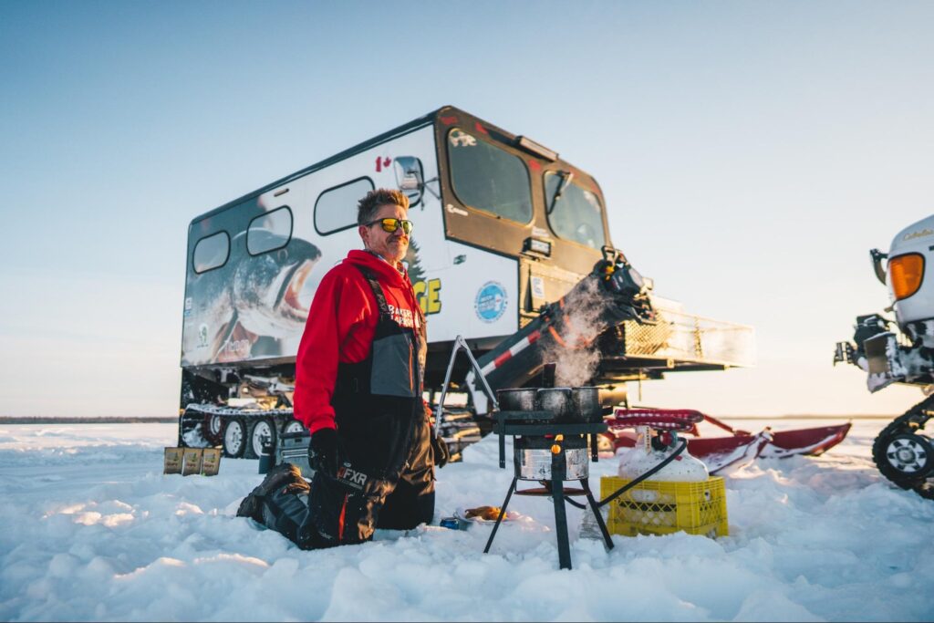 Man cooking on the ice with fishing gear and snow vehicle in the background