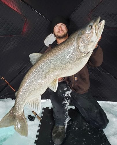 A male ice angler proudly hoists a massive lake trout caught while ice fishing in Manitoba.