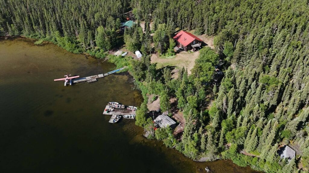 Floatplane tied to the dock at the Kamuchawie Lake fly-in outpost, with fishing boats moored nearby and the red-roofed log lodge in the spruce forest above the shoreline