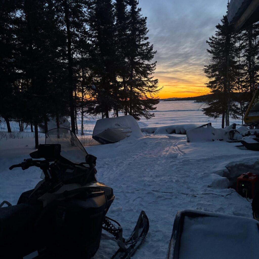 Snowmobile parked at the lodge with sunset over frozen Kamuchawie Lake